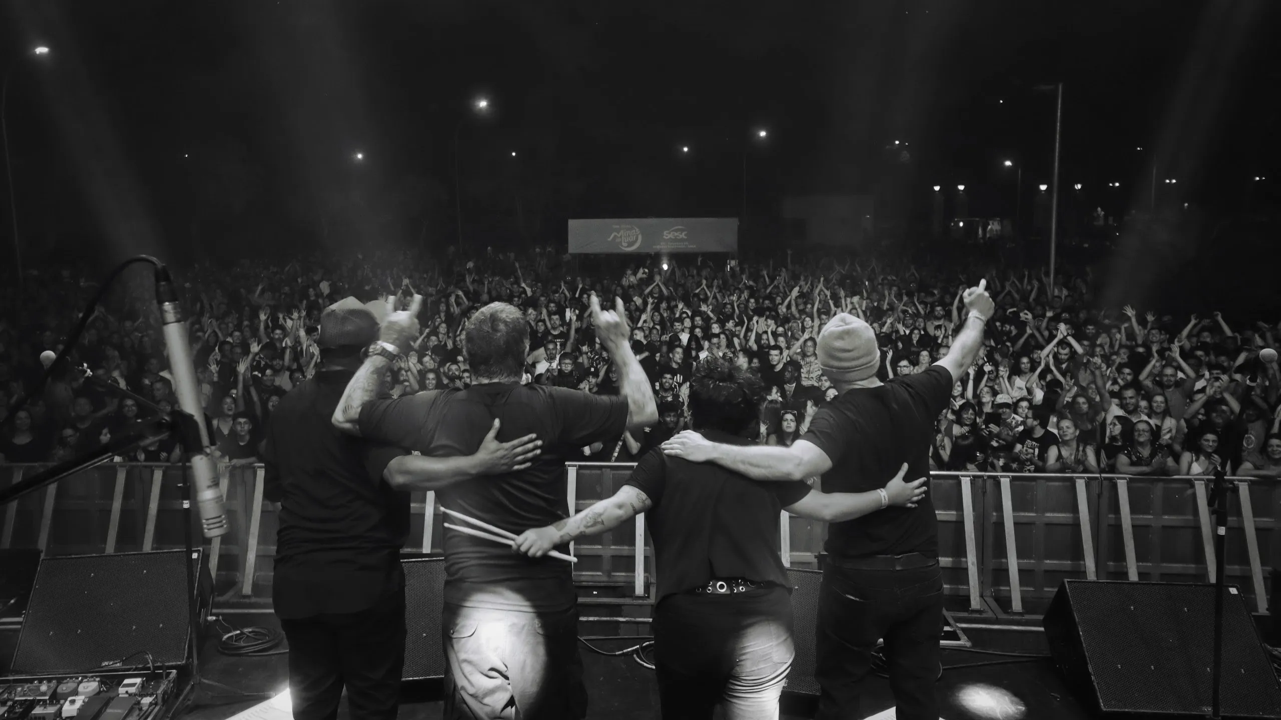 Integrantes da Tianastácia abraçados de costas no palco, em preto e branco, diante de uma plateia grande com os braços erguidos