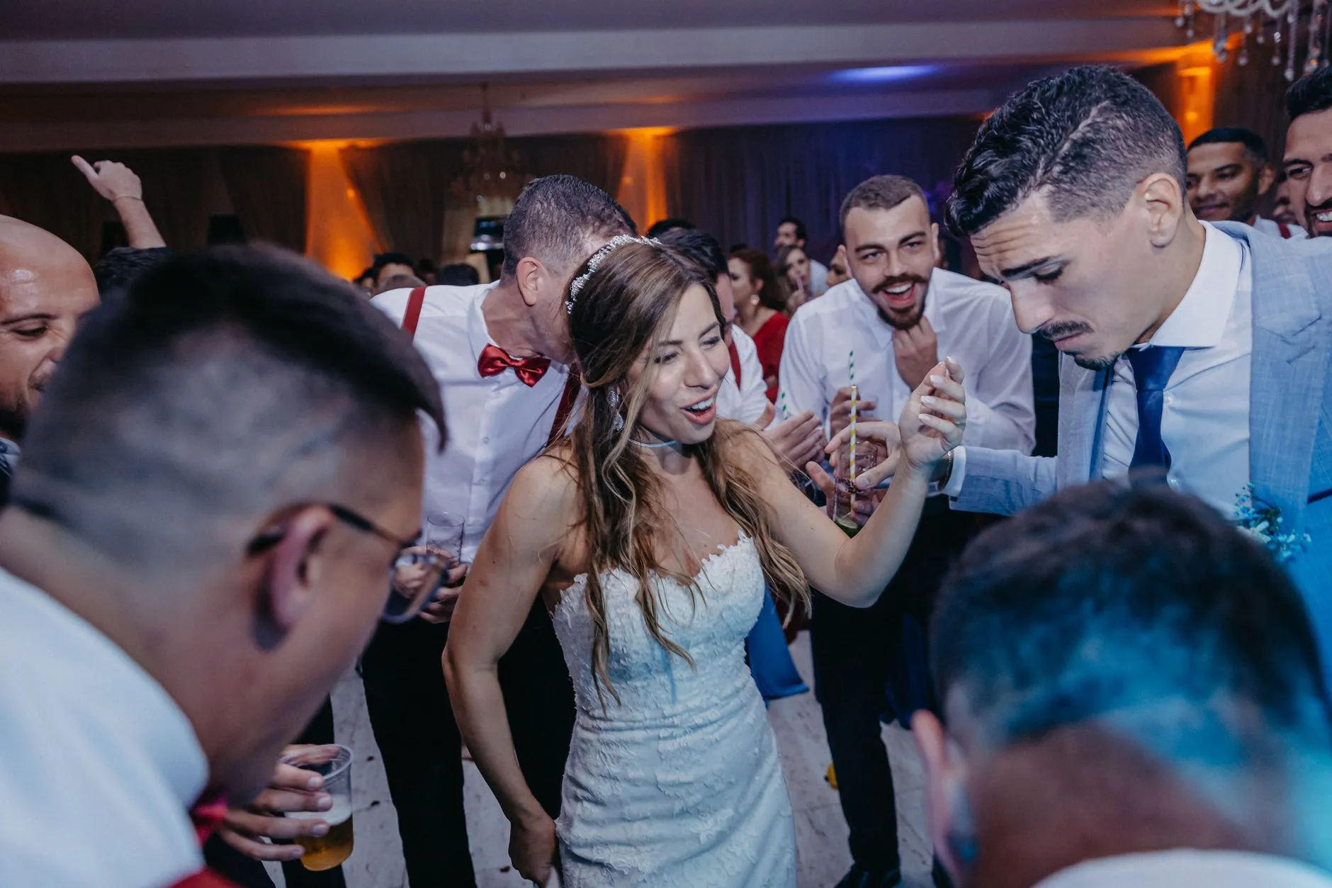 Noiva dançando no meio dos convidados na pista de um salão de casamento, com luzes quentes ao fundo