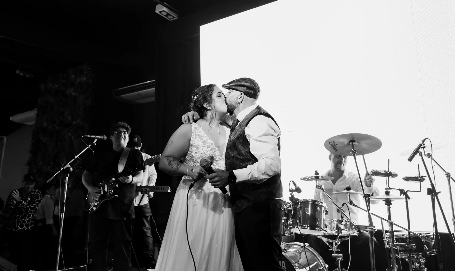 Noivos se beijando no palco durante o show da banda na festa de casamento, com guitarrista e baterista ao fundo