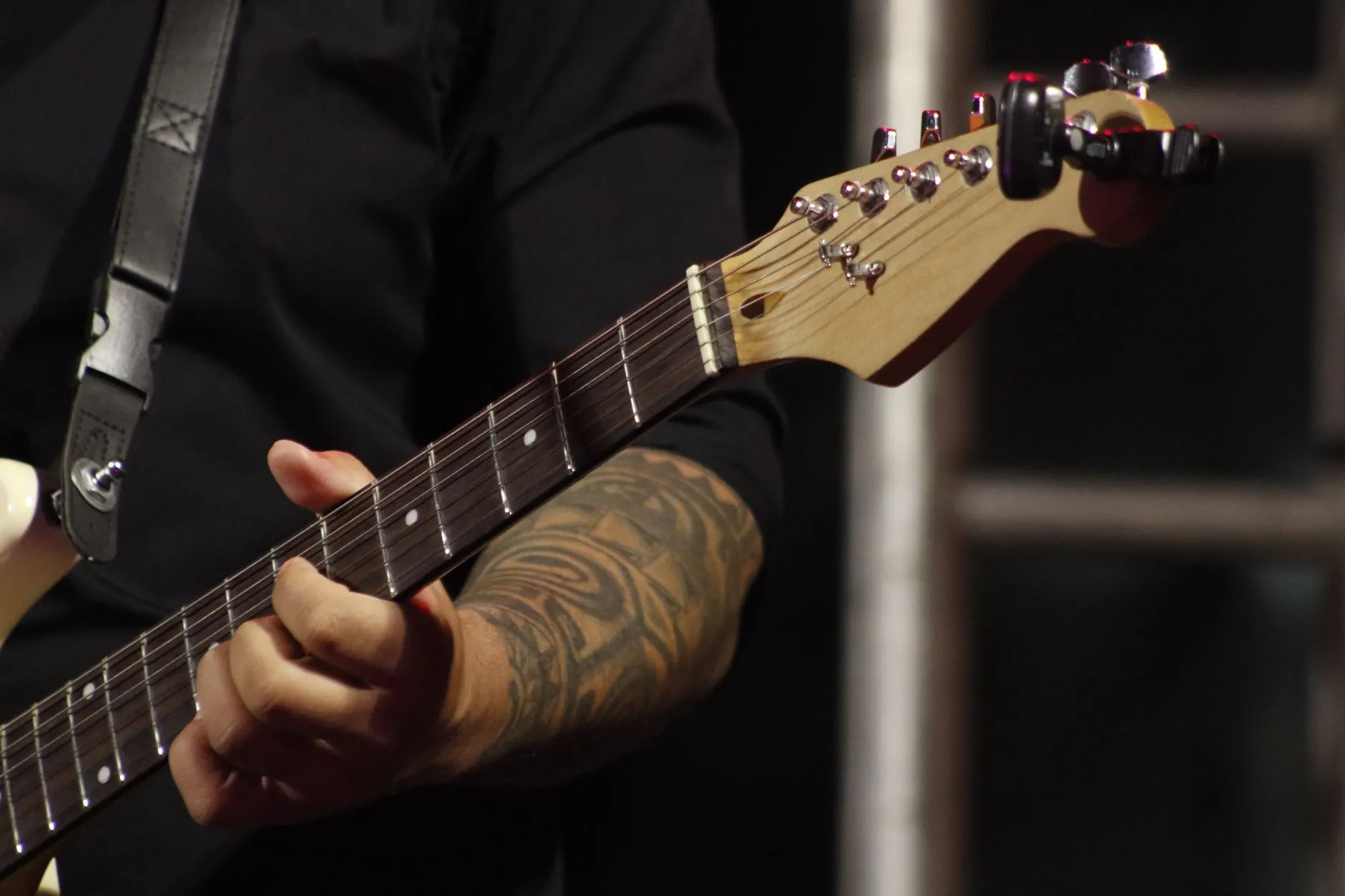 Guitarrista de camisa preta tocando guitarra elétrica no palco, em close no braço do instrumento